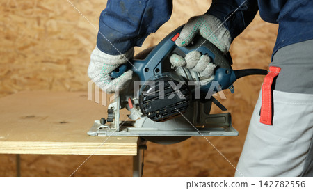 Builder saws a board with a circular saw cutting a wooden plank in a joinery. Carpenter uses portable circular saw tool to cut wood at wooden table in workshop. Worker sawing pine board. Real image Builder saws a board with a circular saw cutting a wooden plank in a joinery. Carpenter uses portable circular saw tool to cut wood at wooden table in workshop. Worker sawing pine board. Real image 142782556