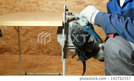 Builder saws a board with a circular saw cutting a wooden plank in a joinery. Carpenter uses portable circular saw tool to cut wood at wooden table in workshop. Worker sawing pine board. Real image Builder saws a board with a circular saw cutting a wooden plank in a joinery. Carpenter uses portable circular saw tool to cut wood at wooden table in workshop. Worker sawing pine board. Real image 142782557