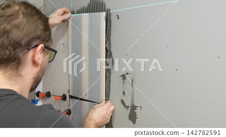 Professional tiler installing ceramic wall tiles with adhesive mortar. Worker hands using leveling system clips. Tiler hand adjusting tile. Wall tiling process. Interior Renovation. Precision Building Professional tiler installing ceramic wall tiles with adhesive mortar. Worker hands using leveling system clips. Tiler hand adjusting tile. Wall tiling process. Interior Renovation. Precision Building 142782591