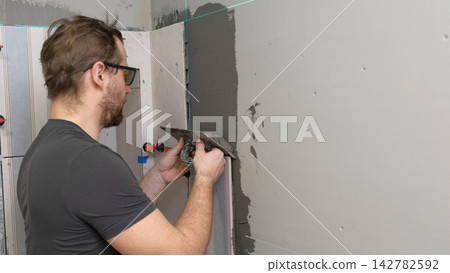 Professional tiler installing ceramic wall tiles with adhesive mortar. Worker hands using leveling system clips. Tiler hand adjusting tile. Wall tiling process. Interior Renovation. Precision Building Professional tiler installing ceramic wall tiles with adhesive mortar. Worker hands using leveling system clips. Tiler hand adjusting tile. Wall tiling process. Interior Renovation. Precision Building 142782592