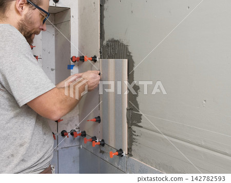 Professional tiler installing ceramic wall tiles with adhesive mortar. Worker hands using leveling system clips. Tiler hand adjusting tile. Wall tiling process. Interior Renovation. Precision Building Professional tiler installing ceramic wall tiles with adhesive mortar. Worker hands using leveling system clips. Tiler hand adjusting tile. Wall tiling process. Interior Renovation. Precision Building 142782593