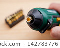 Hex chuck bit holder of green electric screwdriver with blurred set of interchangeable driver tips in the background on a wooden workbench. Home power tools 142783776