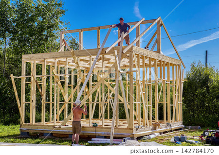 Rafter system installation, young worker positions timber rafters on frame house while second builder assists with roof structure construction. Rafter system installation, young worker positions timber rafters on frame house while second builder assists with roof structure construction. 142784170