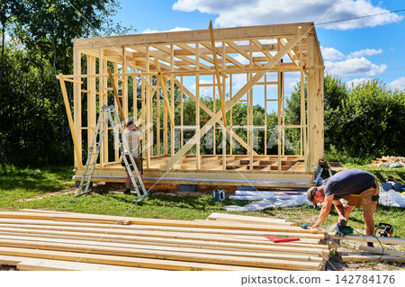 Construction crew of two workers builds timber frame as one cuts boards with circular saw and another fastens braces to wall studs. Construction crew of two workers builds timber frame as one cuts boards with circular saw and another fastens braces to wall studs. 142784176