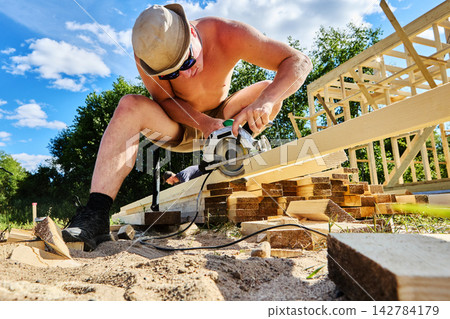 Cutting lumber with circular saw, young worker operates power tool during construction of structural frame. Cutting lumber with circular saw, young worker operates power tool during construction of structural frame. 142784179