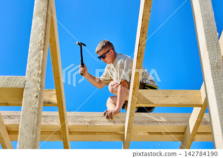 Working at height as young construction worker uses hammer to fasten timber boards on unfinished wooden frame. Working at height as young construction worker uses hammer to fasten timber boards on unfinished wooden frame. 142784190