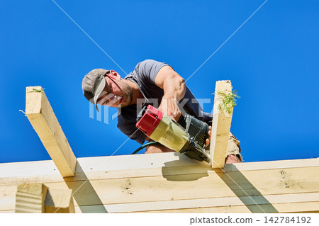 Pneumatic framing nailer drives nails into wooden boards while young worker performs carpentry during timber frame construction. Pneumatic framing nailer drives nails into wooden boards while young worker performs carpentry during timber frame construction. 142784192