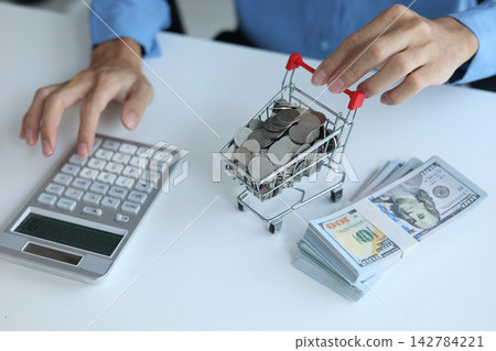 Miniature shopping cart filled with coins and stack of money on white table. Financial planning and E-commerce concept. Miniature shopping cart filled with coins and stack of money on white table. Financial planning and E-commerce concept. 142784221