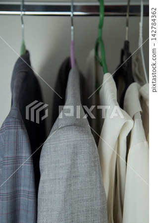 A close-up shows the upper part of a grey melange jacket and adjacent white clothes hanging on a steel rod with multicolored hooks. Concept: emphasis on shoulder geometry, fabric material. High 142784362