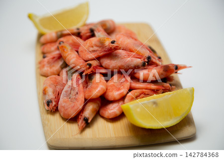 There is a handful of frozen pink shrimp on a light wooden cutting board, with lemon slices diagonally arranged. Concept: diagonal composition on a board. High quality photo There is a handful of frozen pink shrimp on a light wooden cutting board, with lemon slices diagonally arranged. Concept: diagonal composition on a board. High quality photo 142784368