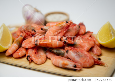 A close-up side view of frozen shrimps on a wooden board, two lemon slices diagonally, a salt shaker and a head of garlic next to it. Concept: an expanded set of seafood seasonings. High quality photo A close-up side view of frozen shrimps on a wooden board, two lemon slices diagonally, a salt shaker and a head of garlic next to it. Concept: an expanded set of seafood seasonings. High quality photo 142784370