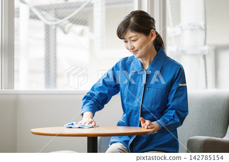A middle-aged cleaning staff member in work clothes wipes down an office table. A middle-aged cleaning staff member in work clothes wipes down an office table. 142785154