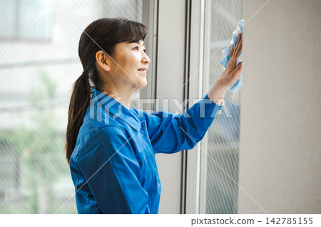 一名身穿工作服的中年清潔工正在擦拭建築物的窗戶。 一名身穿工作服的中年清潔工正在擦拭建築物的窗戶。 142785155