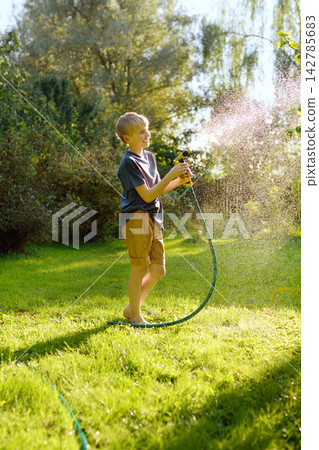 Funny preteen boy pouring cool water on himself and playing with garden hose with sprinkler in sunny backyard. Happy child having fun with water splashes. 142785683