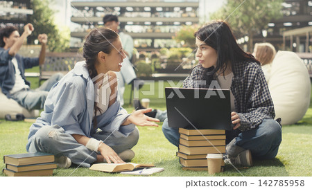 Diverse classmates collaborating on laptop while seated on campus lawn, discussing coursework and taking notes for exam preparation. Peer tutoring and group study. Diverse classmates collaborating on laptop while seated on campus lawn, discussing coursework and taking notes for exam preparation. Peer tutoring and group study. 142785958