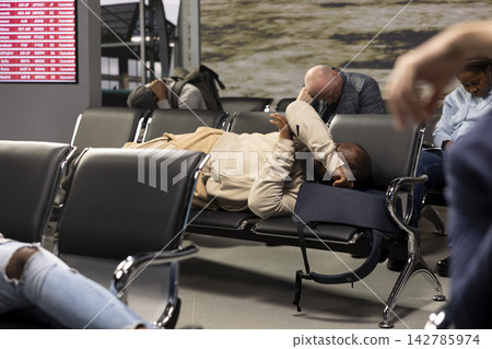 Exhausted man sleeping on metal bench in busy airport lounge, flight delay concept. Tired african american passenger resting during long flight layover, waiting for departure Exhausted man sleeping on metal bench in busy airport lounge, flight delay concept. Tired african american passenger resting during long flight layover, waiting for departure 142785974