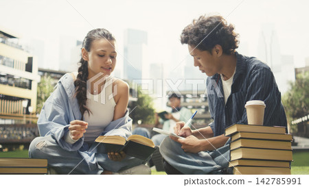 Diverse classmates collaborating with textbooks while seated on campus lawn, discussing coursework and taking notes for exam preparation. Peer tutoring and group study. Diverse classmates collaborating with textbooks while seated on campus lawn, discussing coursework and taking notes for exam preparation. Peer tutoring and group study. 142785991
