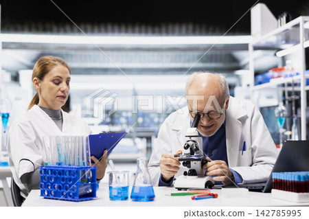 Senior lab manager examining tissue under microscope next to assistant writing findings. Elderly laboratory team leader checking cellular structure integrity next to woman documenting report 142785995