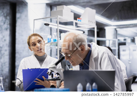 Senior scientist inspecting new drug compound under microscope, helped by assistant reporting findings. Elderly man in research facility verifying medication quality, intern apprentice taking notes 142785997