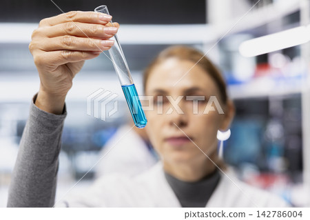 Close up of clinical lab molecular biologist analyzing blue liquid chemical sample properties in glass test tube. Research facility middle aged expert holds vial containing compound for RD study Close up of clinical lab molecular biologist analyzing blue liquid chemical sample properties in glass test tube. Research facility middle aged expert holds vial containing compound for RD study 142786004