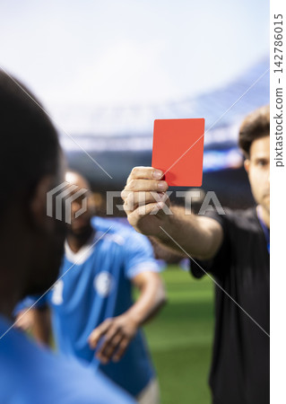 Close-up of red card shown to protesting soccer player during heated match dispute, with teammates complaining about dismissal decision. Referee sanctioning rules violation. Close-up of red card shown to protesting soccer player during heated match dispute, with teammates complaining about dismissal decision. Referee sanctioning rules violation. 142786015