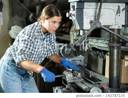 Girl worker at metallurgical plant drills hole in metal plate using drilling machine Girl worker at metallurgical plant drills hole in metal plate using drilling machine 142787135