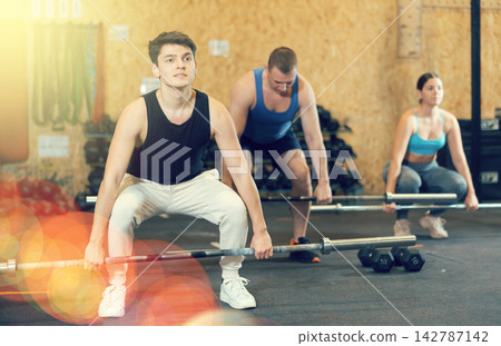 Young male bodybuilder performing strength and endurance exercises during group barbell lifting in CrossFit class 142787142