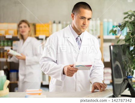 Man pharmacy employee works with computer, girl assistant work in background Man pharmacy employee works with computer, girl assistant work in background 142787342