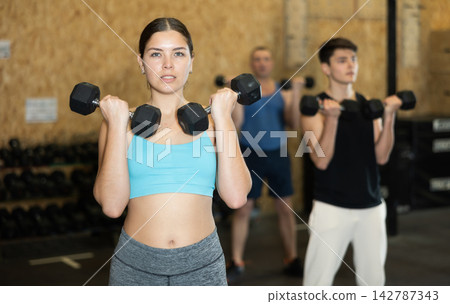 Sportive strong young female in activewear lifting dumbbells during group workout class in gym indoors. Functional training concept Sportive strong young female in activewear lifting dumbbells during group workout class in gym indoors. Functional training concept 142787343