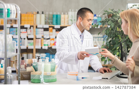 Young male pharmacist offering pills box to client in chemist's shop Young male pharmacist offering pills box to client in chemist's shop 142787384