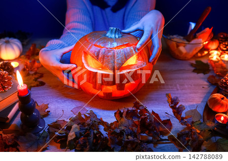 Halloween preparation at home. Hands hold a carved pumpkin decoration for a fun party celebration, keeping the spooky autumn entertainment and tradition alive. Halloween preparation at home. Hands hold a carved pumpkin decoration for a fun party celebration, keeping the spooky autumn entertainment and tradition alive. 142788089