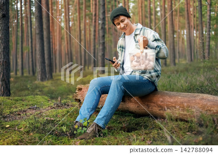 man sitting on a forest log holding the smartphone and a basket full of wild mushrooms, enjoying a seasonal autumn harvest, foraging, and nature connection. man sitting on a forest log holding the smartphone and a basket full of wild mushrooms, enjoying a seasonal autumn harvest, foraging, and nature connection. 142788094