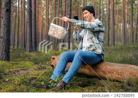 man sitting on a forest log takes a smartphone photo of his autumn harvest. Enjoying outdoor activity and nature connection, he captures his wild foraging basket full of fresh mushrooms. man sitting on a forest log takes a smartphone photo of his autumn harvest. Enjoying outdoor activity and nature connection, he captures his wild foraging basket full of fresh mushrooms. 142788095