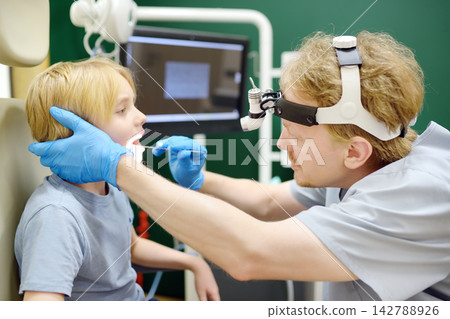 An experienced otolaryngologist examines a patient's throat using a medical tongue depressor in the consulting room. The professional ENT specialist is at their workplace. An experienced otolaryngologist examines a patient's throat using a medical tongue depressor in the consulting room. The professional ENT specialist is at their workplace. 142788926