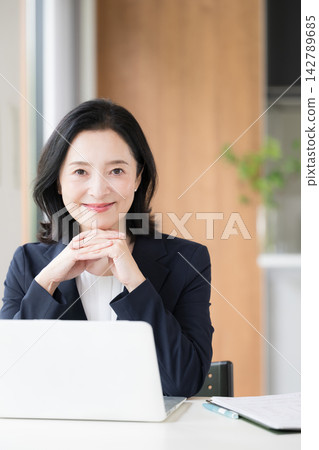 A female office worker working on her laptop; a smiling businesswoman working at her desk in the office, looking directly at the camera. 142789685