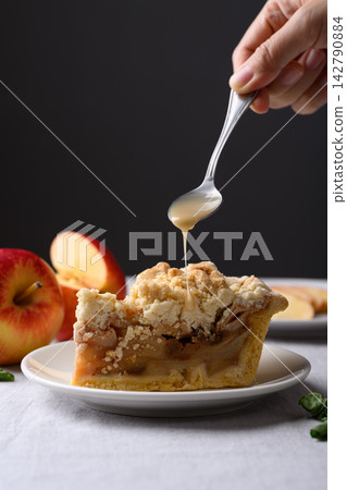 Apple crumble pie slice on a white plate with caramel sauce poured from a spoon Apple crumble pie slice on a white plate with caramel sauce poured from a spoon 142790884
