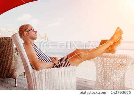 Man relaxing beach sits in a white wicker chair with his feet up on a table at a coastal resort terrace under the sun 142793606