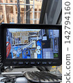The monitor in the crane operator's cab displays the construction site of a wooden house and the work being done from above. 142794160