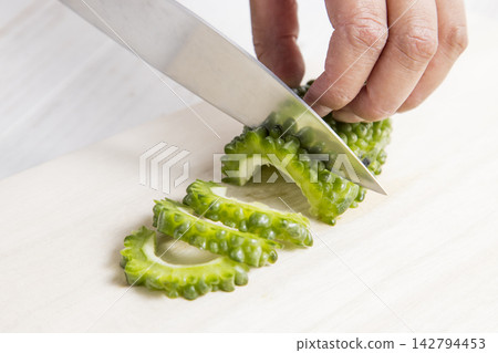 The hands that are slicing the bitter melon, from which the seeds have been removed, into thin slices with a knife. The hands that are slicing the bitter melon, from which the seeds have been removed, into thin slices with a knife. 142794453
