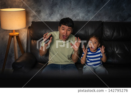 shocked and scared father and daughter watching horror movie tv in the living room at night shocked and scared father and daughter watching horror movie tv in the living room at night 142795276