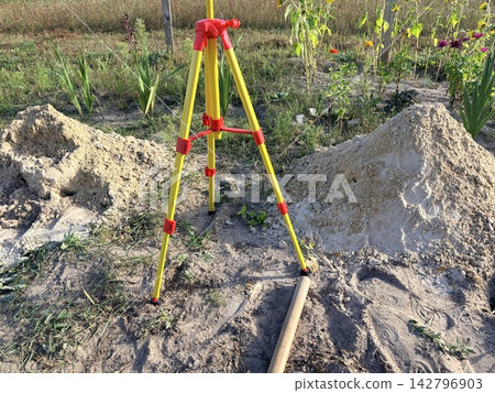 A surveying tripod standing between piles of sand in the garden. site preparation, land surveying,garden renovation. A surveying tripod standing between piles of sand in the garden. site preparation, land surveying,garden renovation. 142796903