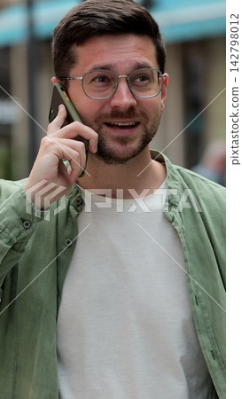 Vertical video. Young man in glasses having phone conversation on the street. Businessman talking on smartphone outdoors. Gadget usage concept. Vertical video. Young man in glasses having phone conversation on the street. Businessman talking on smartphone outdoors. Gadget usage concept. 142798012