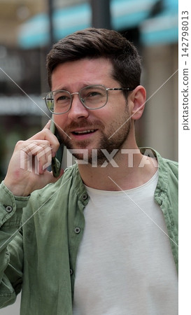 Vertical video. Young man in glasses having phone conversation on the street. Businessman talking on smartphone outdoors. Gadget usage concept. Vertical video. Young man in glasses having phone conversation on the street. Businessman talking on smartphone outdoors. Gadget usage concept. 142798013
