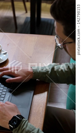 Vertical video. Top view of man in casual clothes is typing on a laptop, sitting at a table. Student is studying in cafe at table with coffee cup. People and technology concept. Vertical video. Top view of man in casual clothes is typing on a laptop, sitting at a table. Student is studying in cafe at table with coffee cup. People and technology concept. 142798153