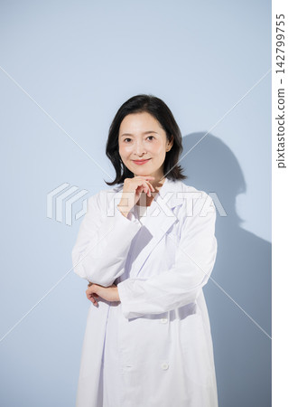 一張日本女醫生的肖像,她身穿白大褂,擺出沉思的姿勢,對著鏡頭微笑。 一張日本女醫生的肖像,她身穿白大褂,擺出沉思的姿勢,對著鏡頭微笑。 142799755