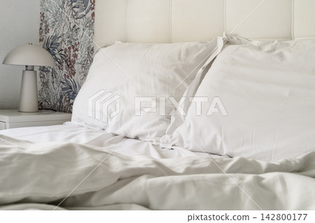 Cozy bedroom with white bedding and a bedside lamp in a quiet space Cozy bedroom with white bedding and a bedside lamp in a quiet space 142800177