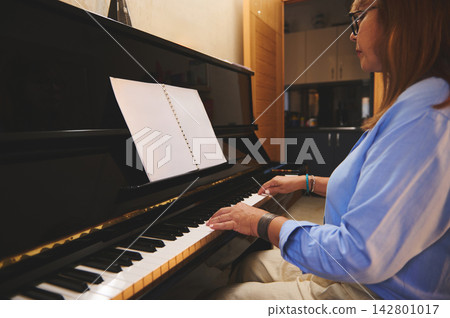 Woman playing piano at home with open sheet music and focused expression Woman playing piano at home with open sheet music and focused expression 142801017