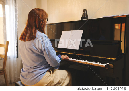 Woman playing upright piano at home with sheet music, practicing and reading notes 142801018