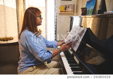Middle-aged woman practicing piano with sheet music in a cozy sunlit living room Middle-aged woman practicing piano with sheet music in a cozy sunlit living room 142801019