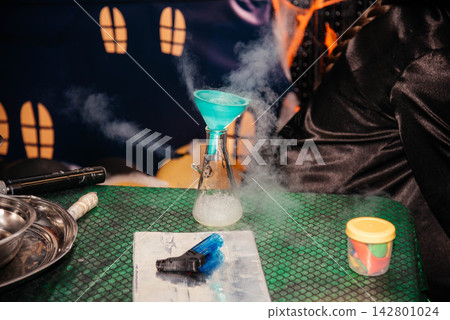 Person in wizard black robe pouring liquid nitrogen from a large dewar tank into a glass flask using a funnel during a scientific show or Halloween theme party. Steaming reaction experiment with smoke 142801024